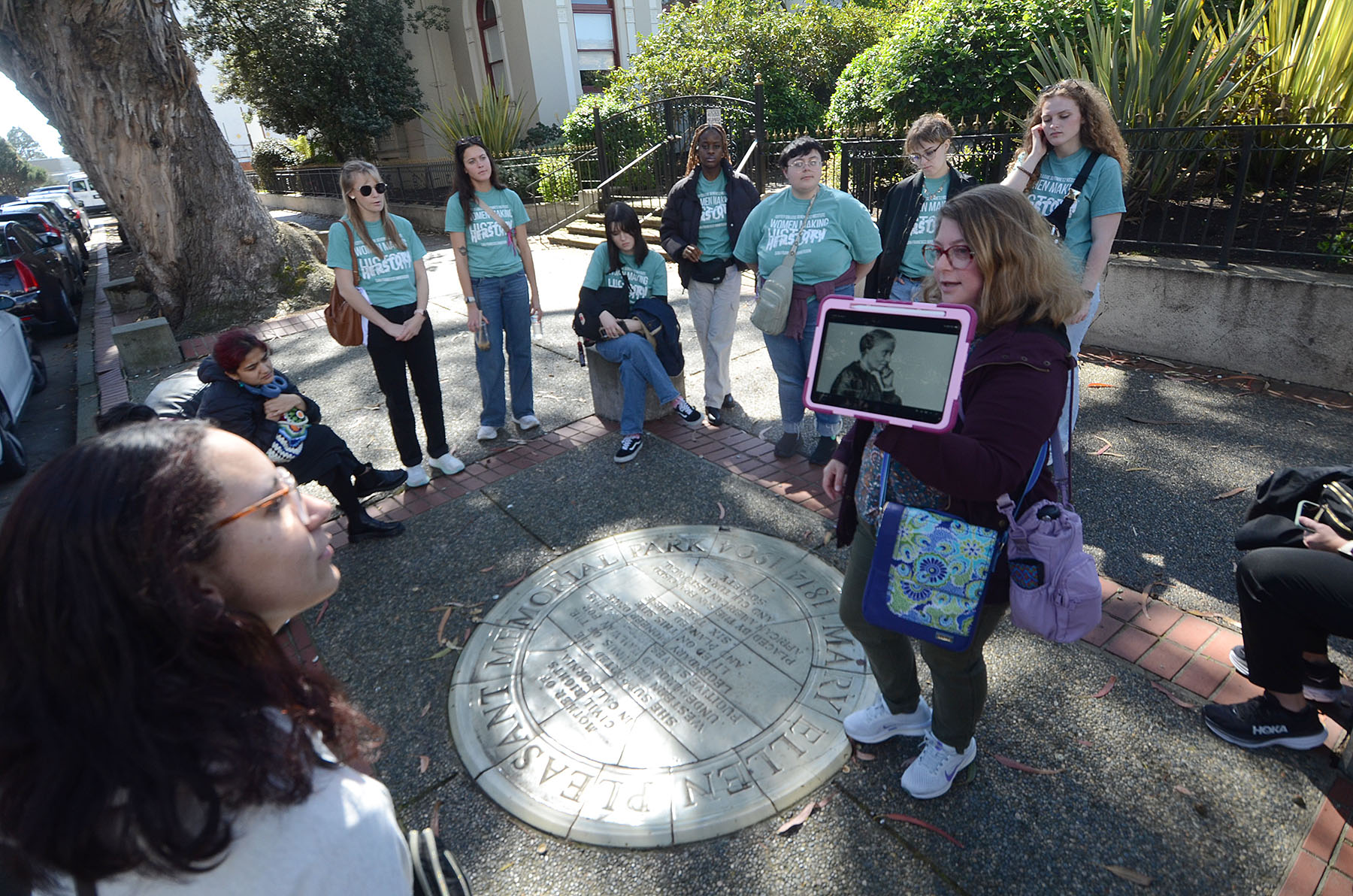 Tour guide shows a historical photo to a group of students gathered around a circular plaque at Mary Ellen Pleasant Memorial Park in San Francisco.